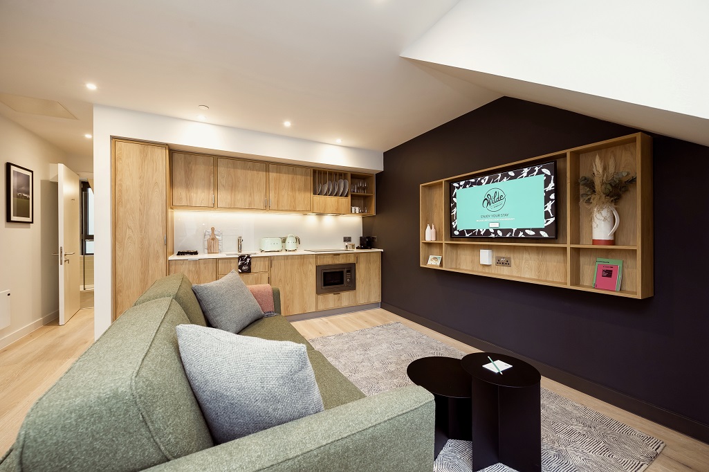A modern living room with a large green sofa opposite a black wall with a television screen attached